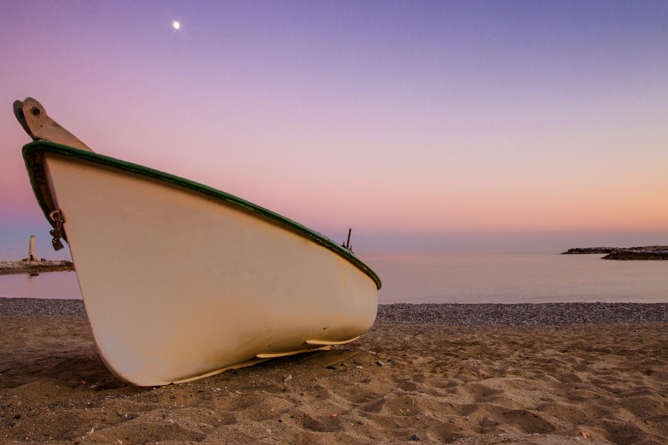 A white wooden rowing boat pulled up onto the sand with a lighthouse behind it on a beach at Puerto Banús in Malaga province Spain. Sky is at sunset and is a pink purple colour with the moon in the distance