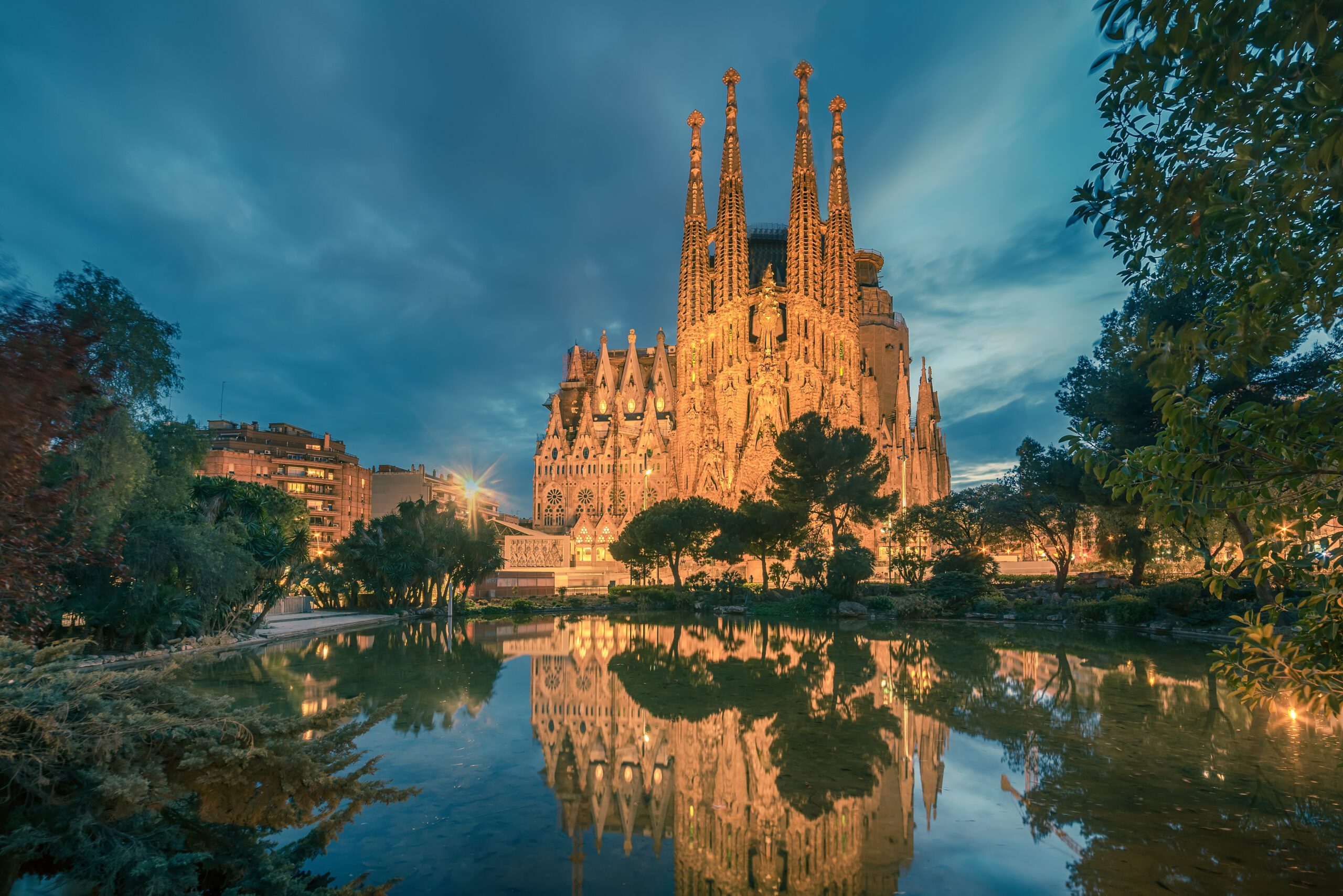 The Sagrada Familia lit up at night with a lake and trees located in front of it.