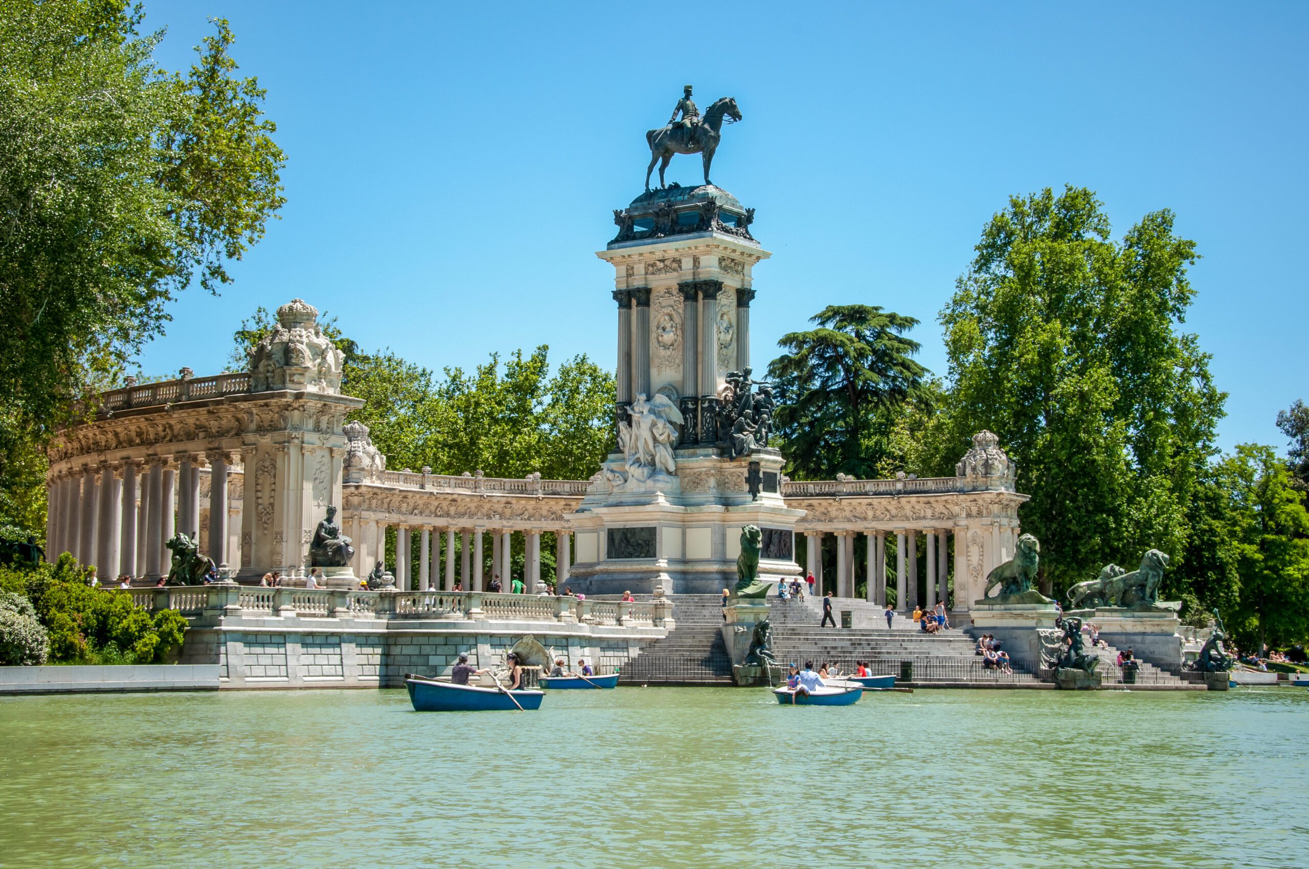 Image shows The main park of the city of Madrid - Buen Retiro Park - there is a river with boats on and historical buildings and a statue with a horse and rider on top.