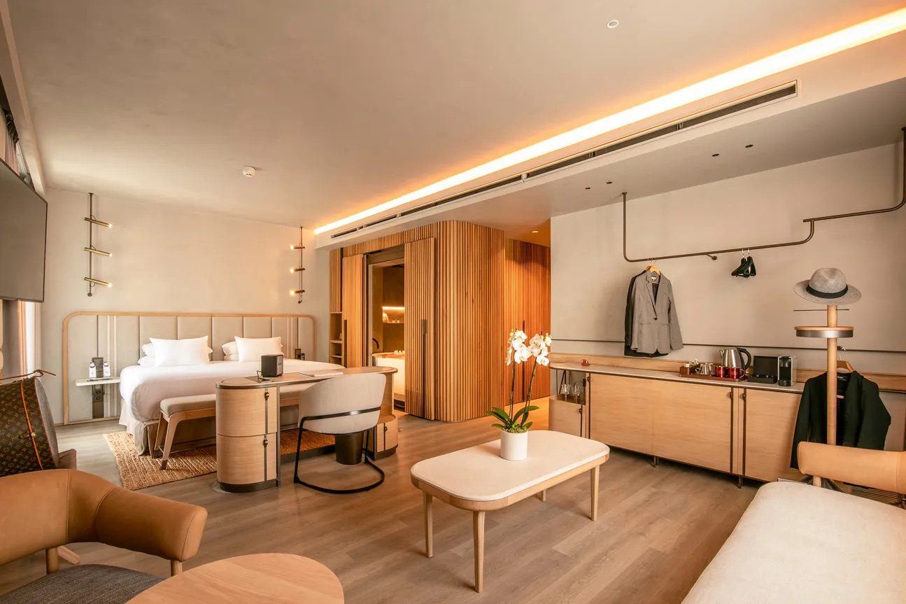 A spacious bedroom at the luxury hotel, featuring a large bed with pristine white bedsheets, a desk, seating, tables and doors leading into the bathroom.