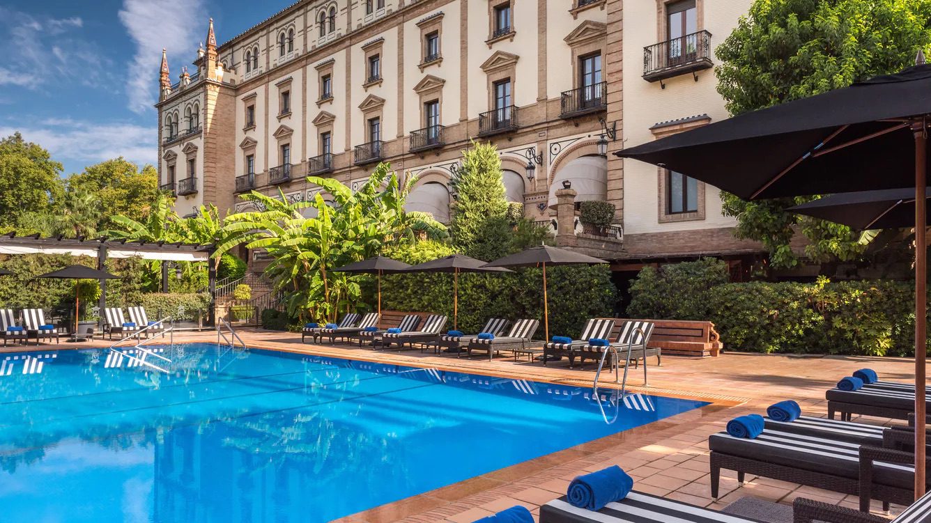 A spacious pool outside the luxury Hotel Alfonso XIII with an array of sun loungers and umbrellas along the edge of pool, surrounded by greenery.