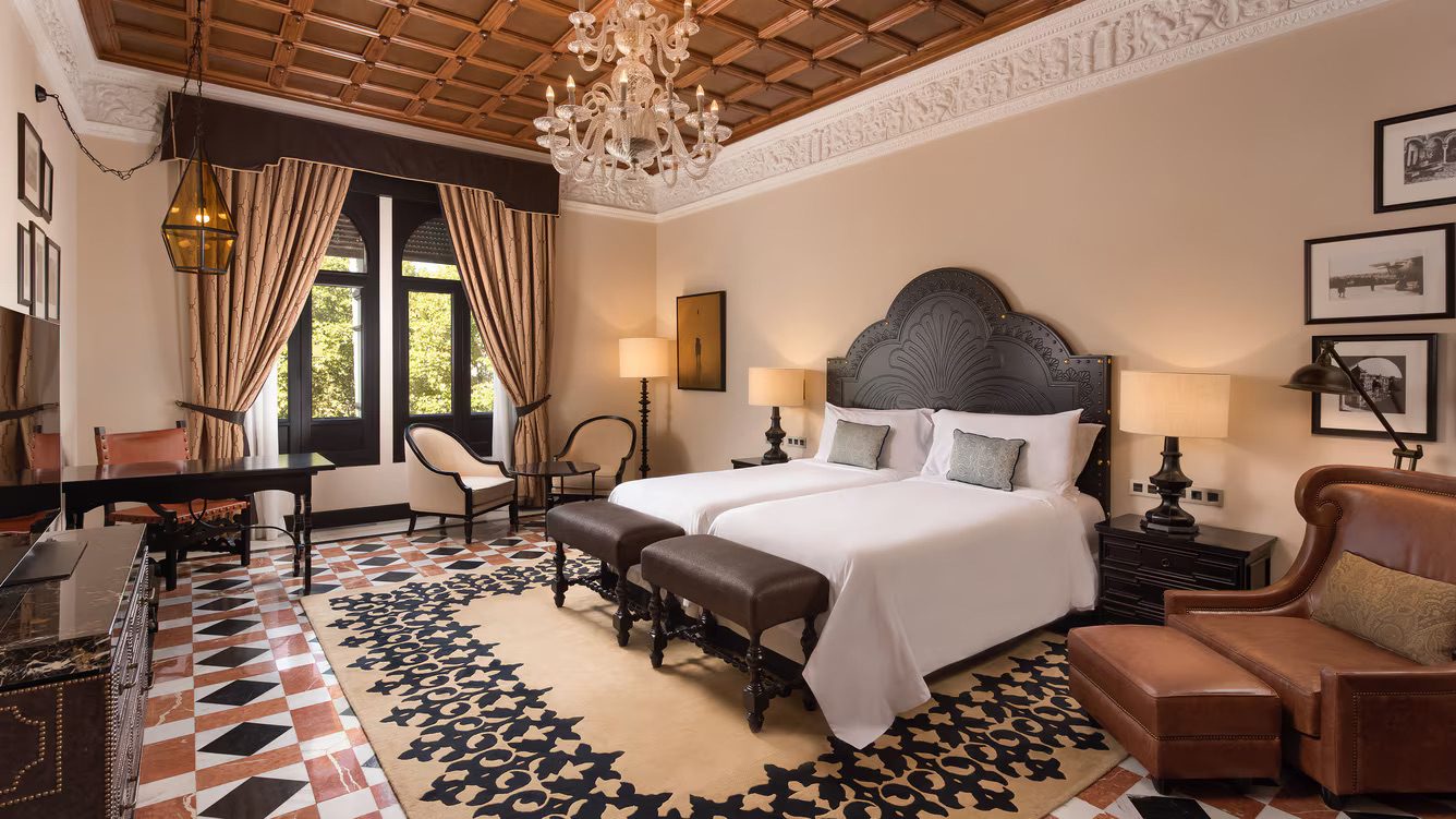 A spacious bedroom in the luxury Hotel Alfonso XIII, featuring a twin bed, armchair, desk, table and chairs and a TV.