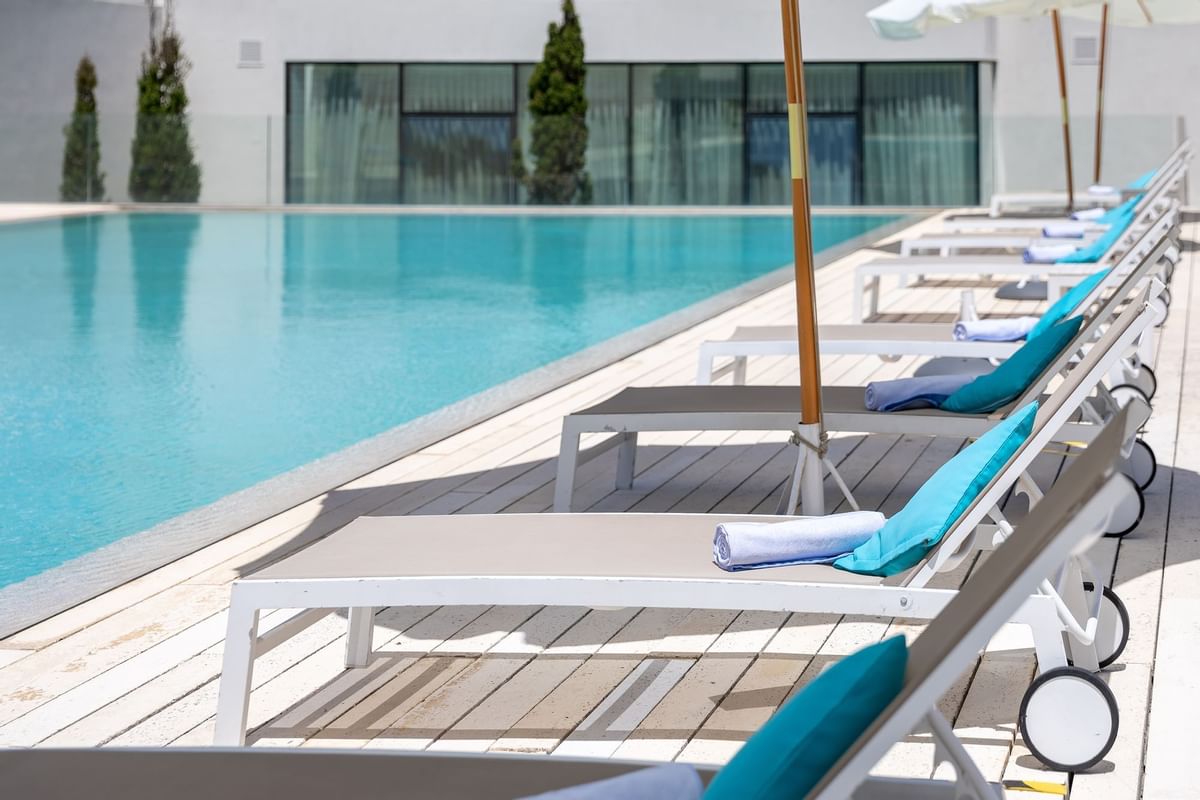 A large outdoor pool with a row of sun loungers lining to pools edge, complete with umbrellas and towels to allow for comfort and shade.