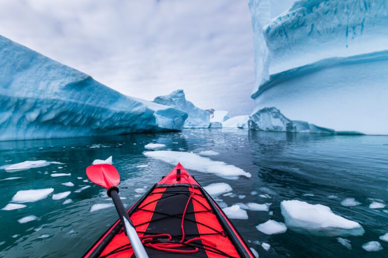 A red inflatable Kayak with an oar across its front in Antarctica between icebergs with ice floating on thw ater surface.