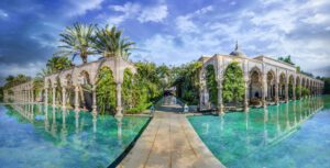 The entrance to a luxury hotel with marble pathways and turquoise decorative ponds eithe side leading to ornate marble archaways to the left and right with climbing plants with pink flowers and palm trees.