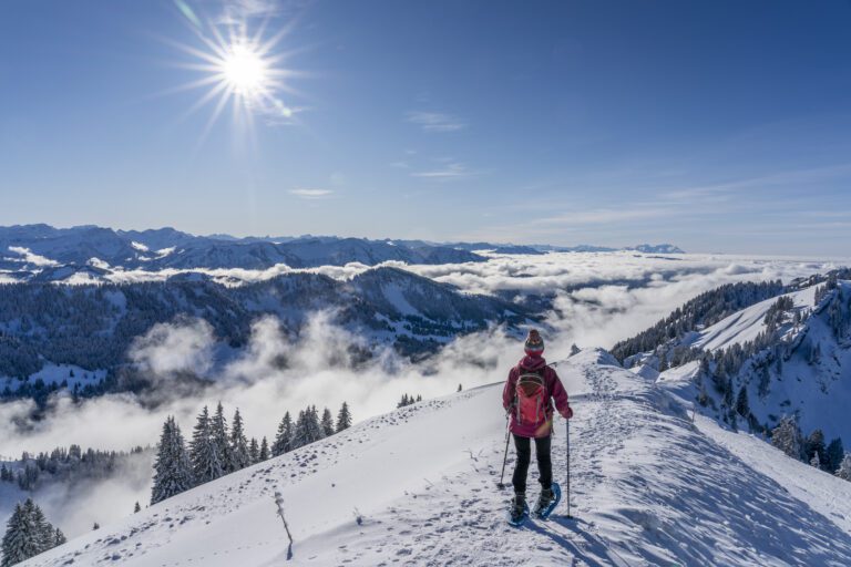 Woman snowshoeing along a ridge with a sea fog below her with the caps of a mountain range poking throug. Stunning blue sky with bright sunlight.