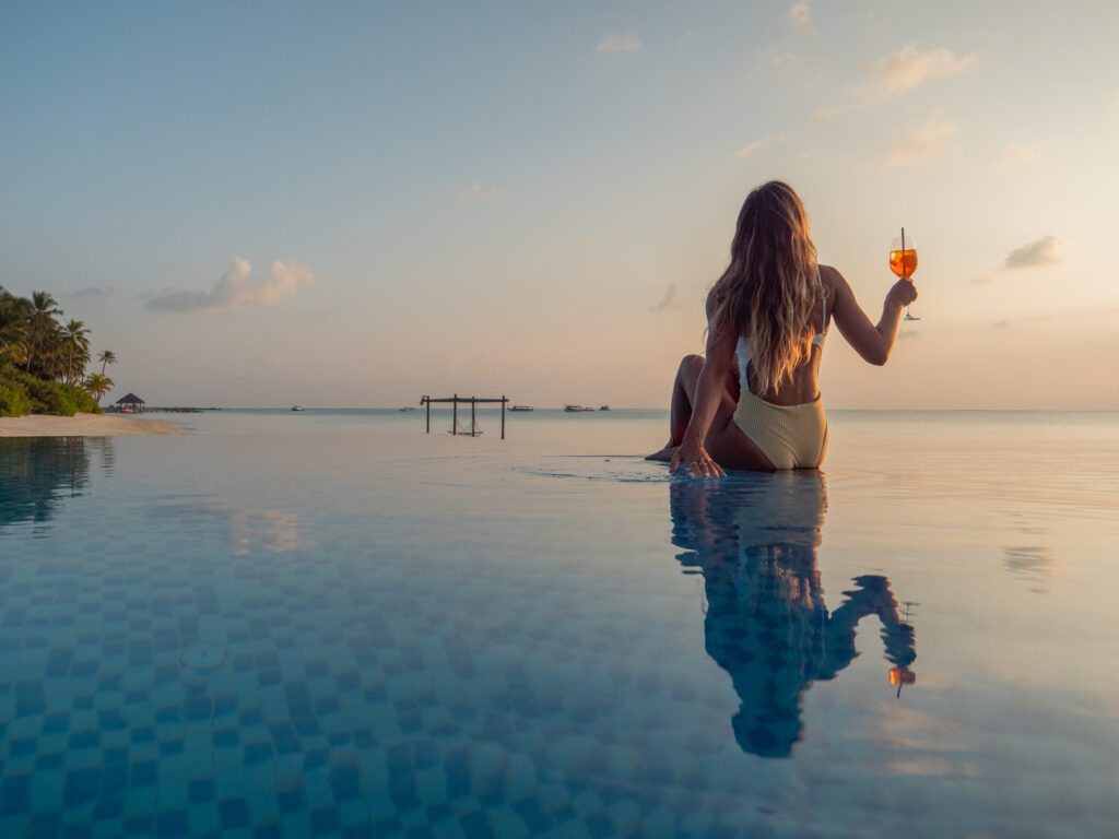 Woman travelling alone, no longer alone in her hotel room, enjoys a drink at sunset on the edge of a swimming pool whilst on a luxury vacation.