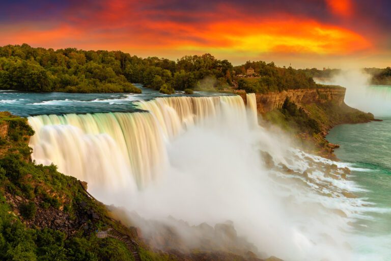 Majestic view of American side of Niagara falls at sunset with cloudy sky lit up red and deep purple and the water crashing over the falls to the rocks below