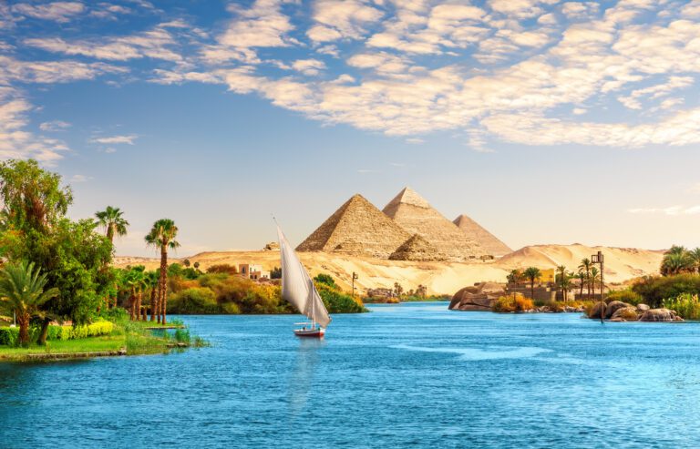 Beautiful Nile scenery with sailboat in the Nile with lush green banks on the left and the arid dry view of the pyramids, Aswan, Egypt.