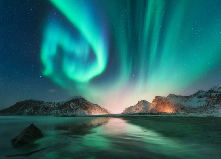 Image of the Aurora Borealis as bright fluorescent greeny blue in swirls over the dark night sky and reflected in the water below in Lofoten Island in Norway