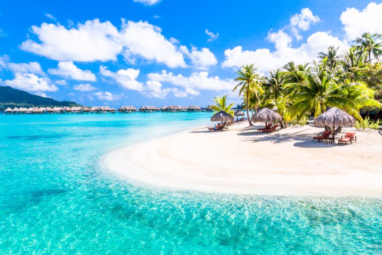 Tropical Beach with white sands, crystal clear turquise sea, palm trees, luxury beach bungalows on stilts in the backgroound under a blue sky with fluffy white clouds