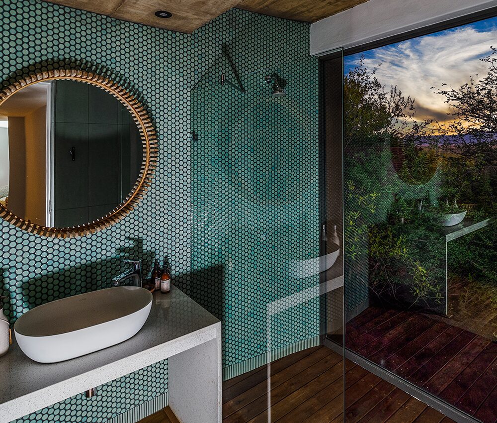 A bathroom at the spa with a sink, mirror and walk-in shower with views of the safari outside the large window.