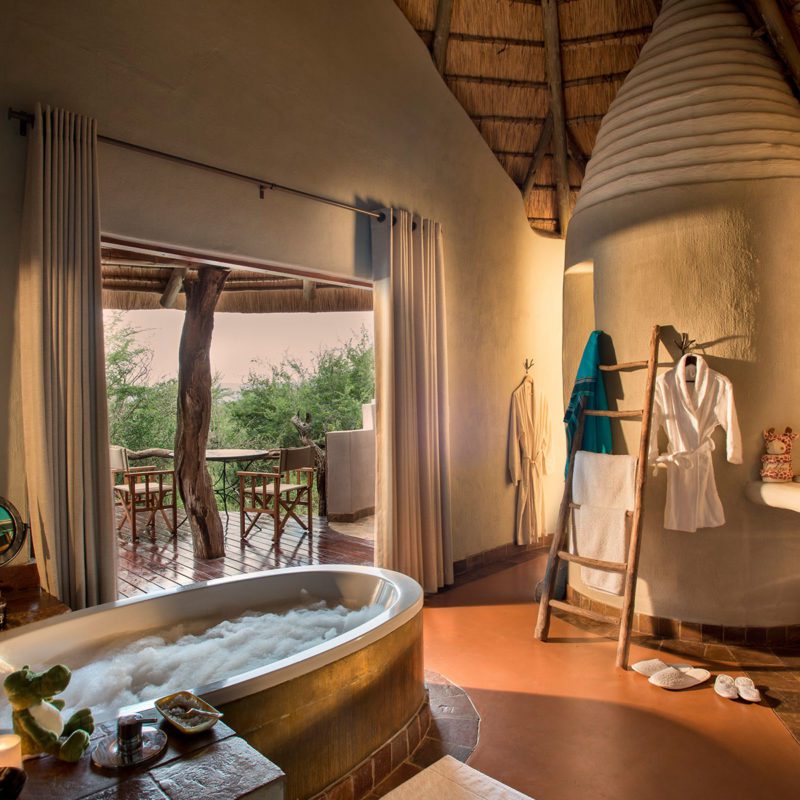 A luxury bathroom with a large bath, robes and towels hanging up and a window looking out onto the private seating area outside.