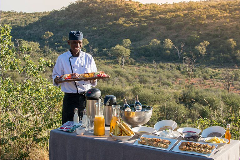 A man serving breakfast outside surrounded by the African greenery.