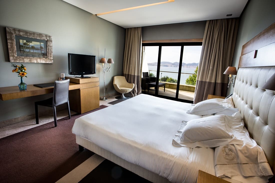 A bedroom in the luxury hotel with a large bed, desk, TV, chairs and large glass doors opening onto a private balcony.
