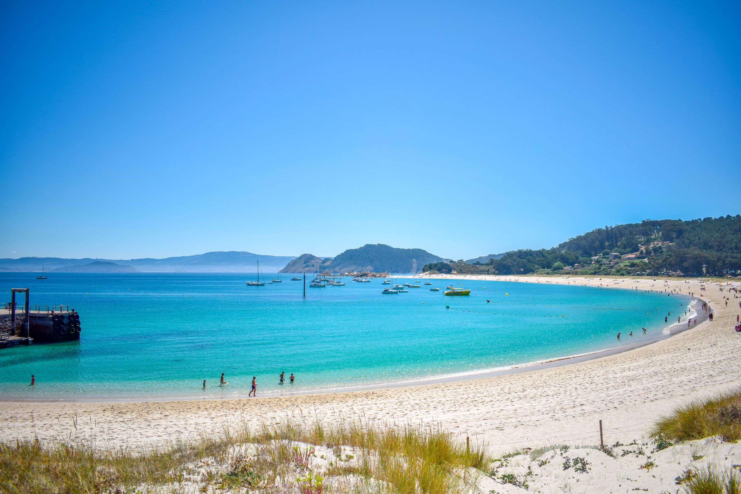 A quiet beach on Cies Islands with sand and bright blue waters and hills in the background.