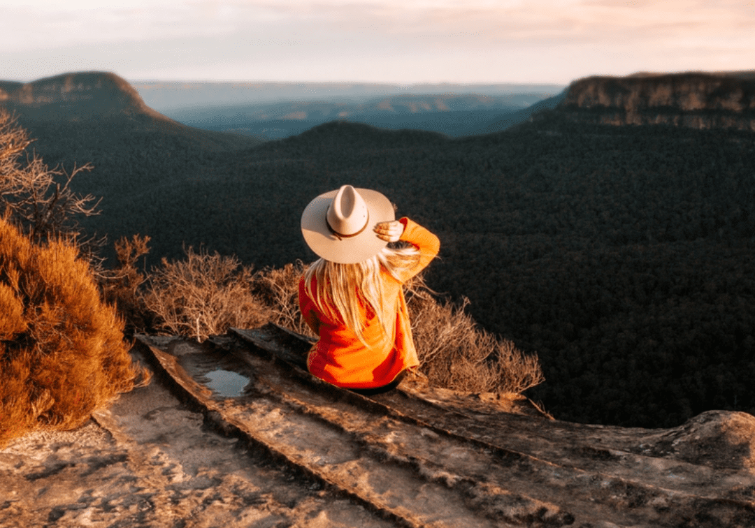 Solo female traveller sat on top of a mountain looking out at the sunset holding her hat embracing her wanderlust.