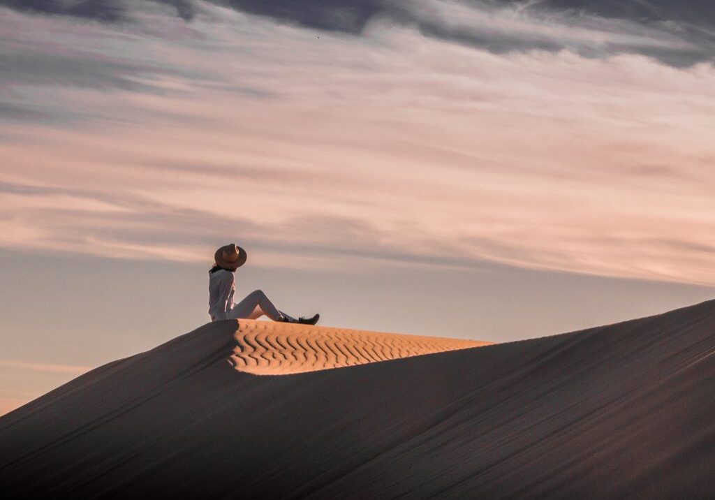 A single Woman, wearing White and a Hat, sitting on top of a huge Sand Dune, looking at the Dramatic Colorful Sky.